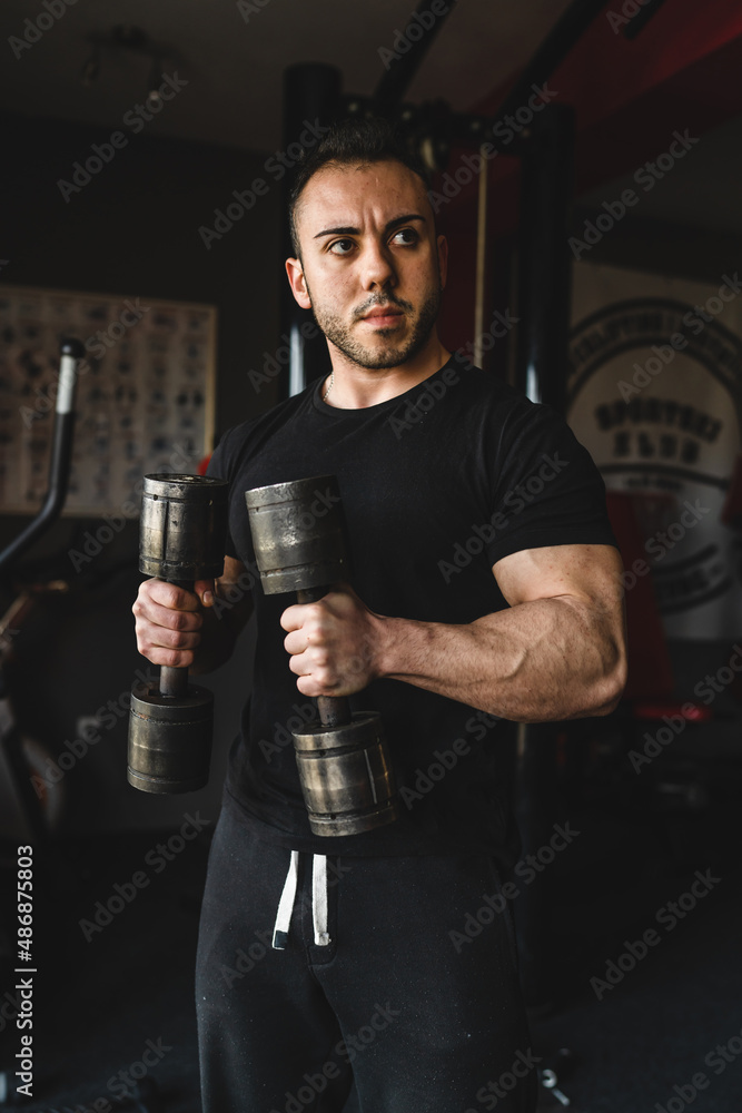 One man young adult caucasian male bodybuilder training arms bicep flexing muscles with dumbbell while standing in the gym wearing black shirt dark photo real people copy space front view waist up One man young adult caucasian male bodybuilder training arms bicep flexing muscles with dumbbell while standing in the gym wearing black shirt dark photo real people copy space front view waist up