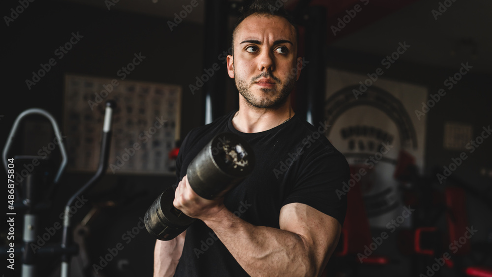 One man young adult caucasian male bodybuilder training arms bicep flexing muscles with dumbbell while standing in the gym wearing black shirt dark photo real people copy space front view waist up One man young adult caucasian male bodybuilder training arms bicep flexing muscles with dumbbell while standing in the gym wearing black shirt dark photo real people copy space front view waist up