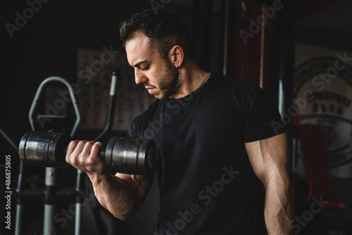One man young adult caucasian male bodybuilder training arms bicep flexing muscles with dumbbell while standing in the gym wearing black shirt dark photo real people copy space front view waist up
