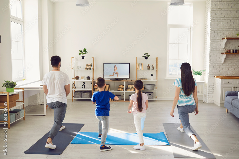 Family having fitness workout at home. Back view of happy mum, dad and kids standing on rubber mats together, watching video workout with gym instructor on TV screen and doing leg stretching exercise Family having fitness workout at home. Back view of happy mum, dad and kids standing on rubber mats together, watching video workout with gym instructor on TV screen and doing leg stretching exercise