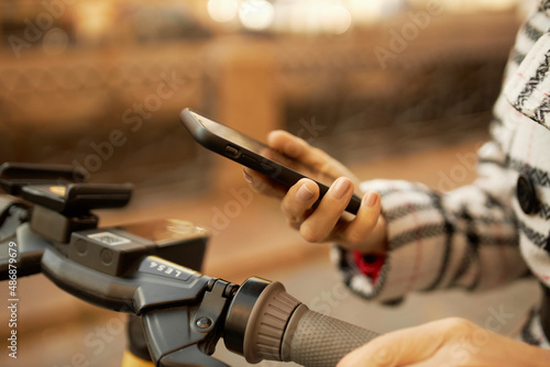 Crop shot of woman hand with manicured nails holding smartphone using application for renting e-scooter. Human and technology. Eco-friendly transport. Big city life. Traveling and adventures