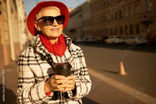 Pretty woman walking down city center streets on sunny Sunday morning holding plastic cup of hot beverage, coffee or tea in hand, wearing warm spring coat and red hat, enjoying moment, smiling