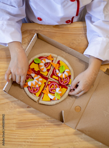 Small boy of Caucasian European appearance in a white chef's cap and a white apron plays toy pizza. Flat lay