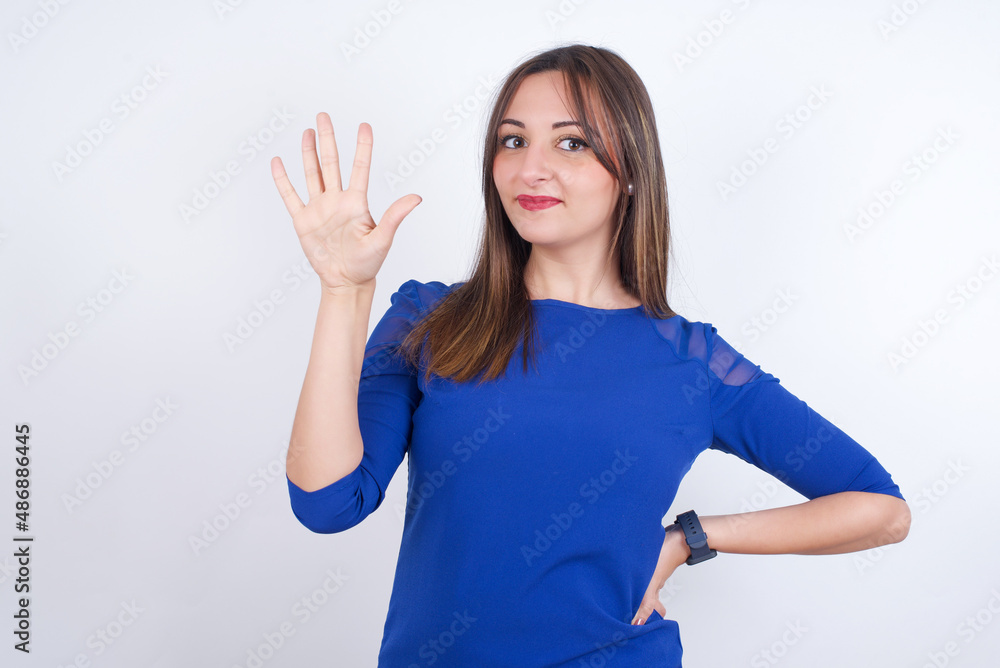 Fototapeta premium Young arab woman wearing blue dress over white backgtound showing and pointing up with fingers number five while smiling confident and happy.