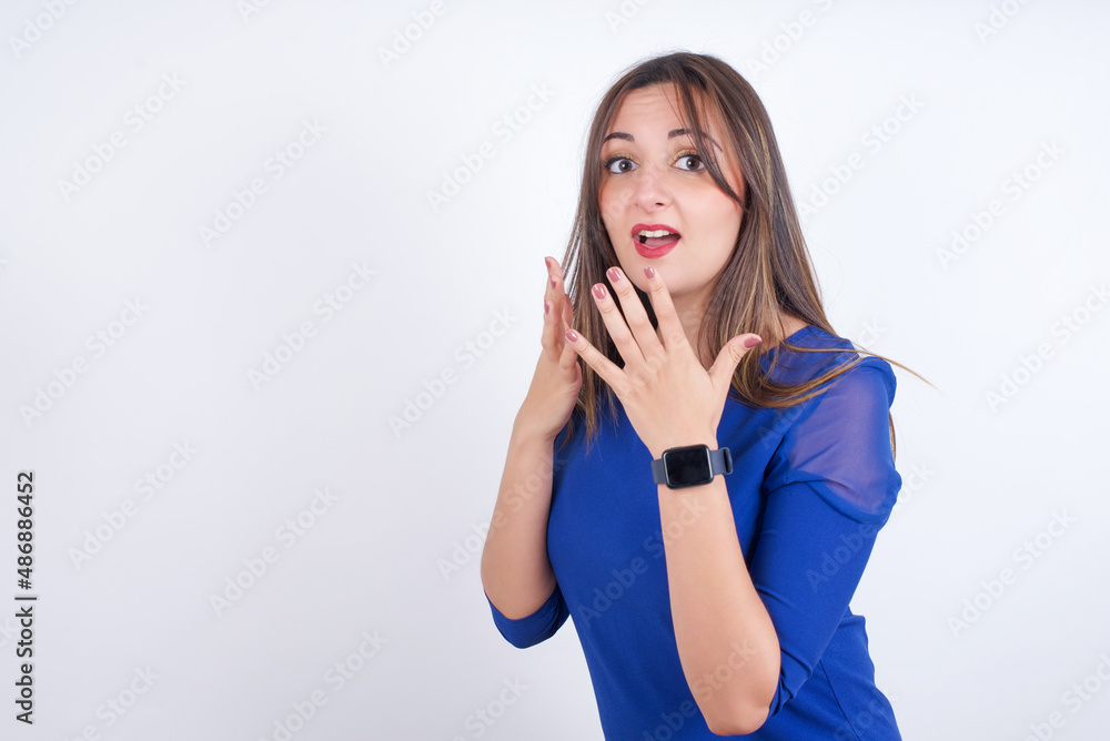 Fototapeta premium Young arab woman wearing blue dress over white backgtound with shocked facial expression, holding hands near face, screaming and looking sideways at something amazing.