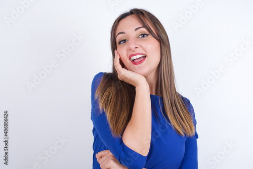 Shocked, astonished Young arab woman wearing blue dress over white backgtound looking surprised in full disbelief wide open mouth with hand near face. Positive emotion facial expression body language.