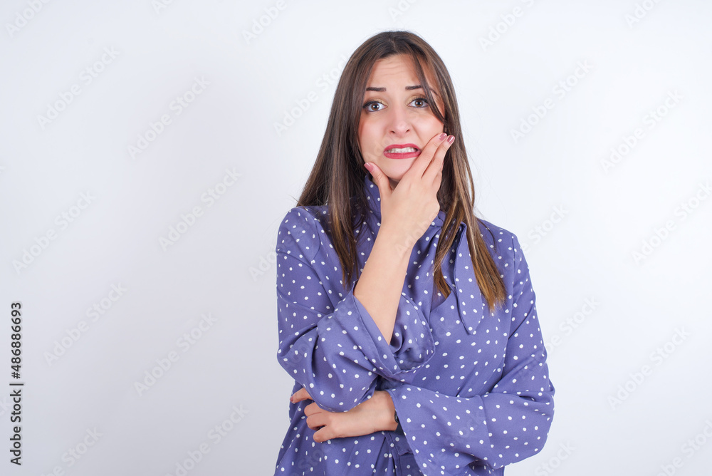 Young arab woman wearing polka-dot clothes over white backgtound covering mouth with hands scared from something or someone bitting nails Young arab woman wearing polka-dot clothes over white backgtound covering mouth with hands scared from something or someone bitting nails