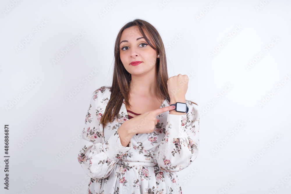 Young arab woman wearing floral shirt over white backgtound in hurry pointing to watch time, impatience, upset and angry for deadline delay