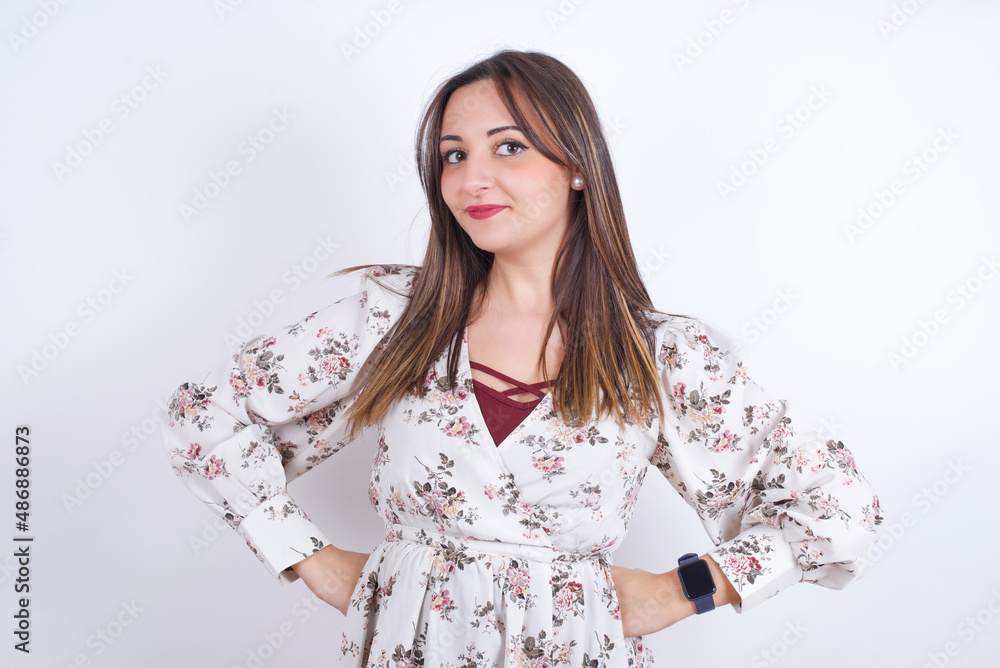 Funny frustrated Young arab woman wearing floral shirt over white backgtound holding hands on waist and silly looking at awkward situation. Funny frustrated Young arab woman wearing floral shirt over white backgtound holding hands on waist and silly looking at awkward situation.