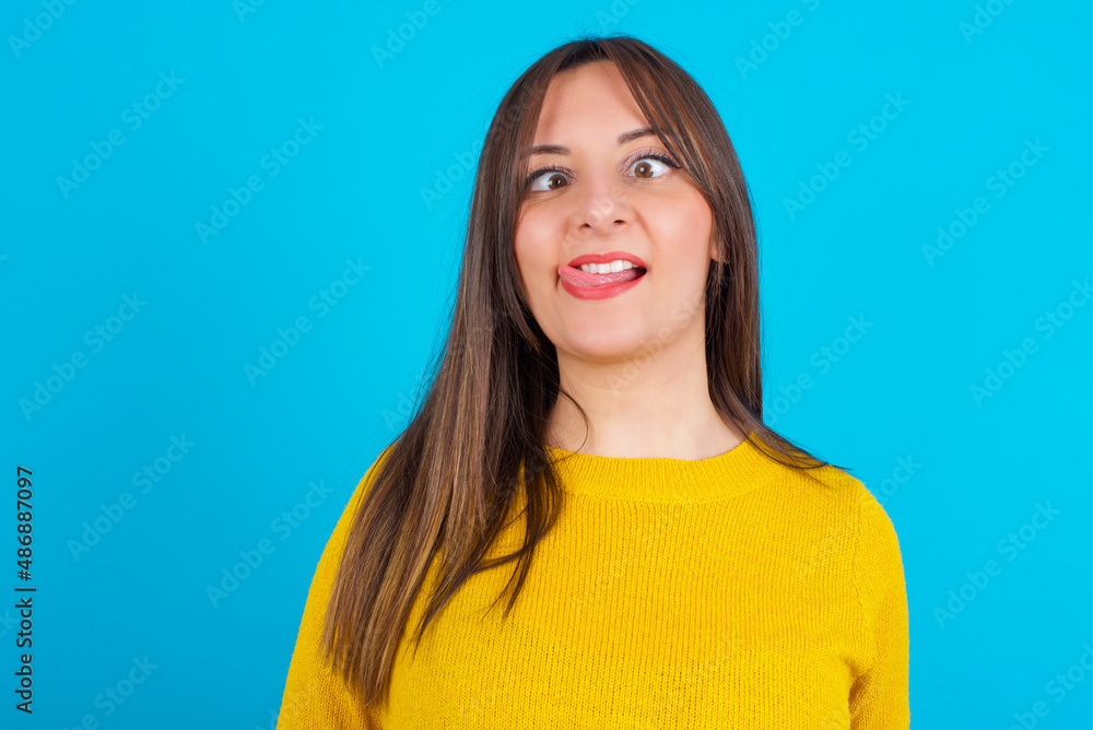 Funny Young arab woman wearing knitted sweater over blue backgtound makes grimace and crosses eyes plays fool has fun alone sticks out tongue. Funny Young arab woman wearing knitted sweater over blue backgtound makes grimace and crosses eyes plays fool has fun alone sticks out tongue.