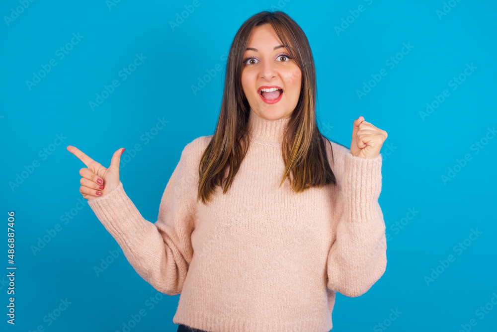 Young arab woman wearing knitted sweater over blue backgtound points at empty space holding fist up, winner gesture. Young arab woman wearing knitted sweater over blue backgtound points at empty space holding fist up, winner gesture.