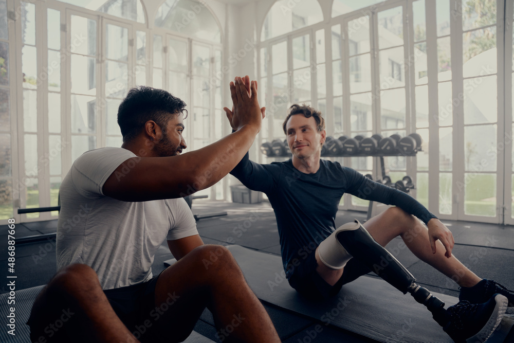 Two friends training in the gym - one with a prosthesis . Two men high fiving in the gym after a good training session Two friends training in the gym - one with a prosthesis . Two men high fiving in the gym after a good training session