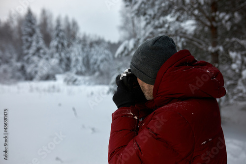 Back view of man taking picture of winter landscape on vintage camera, walking in forest or park, hiking, traveling alone, wearing red coat, grey hat and black gloves. Beauty of nature