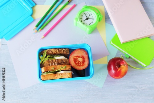 Lunchbox with cereal bread sandwich with cheese, organic vegetables and micro greens, apple, stationery and alarm clock on wooden table, top view