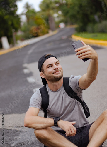 Happy young man backpacker in cap sitting on the middle road and taking a selfie. Travel blogger taking online stream during his hiking travel. 