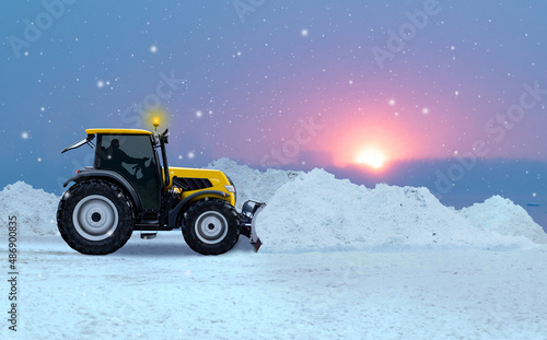 A yellow snowplow road maintenance tractor between piles of snow cleaning a street during heavy snowfall in winter with sunset and copy space in the background