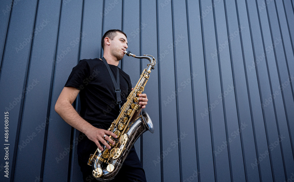 Fototapeta premium Young street musician playing saxophone near the big blue wall