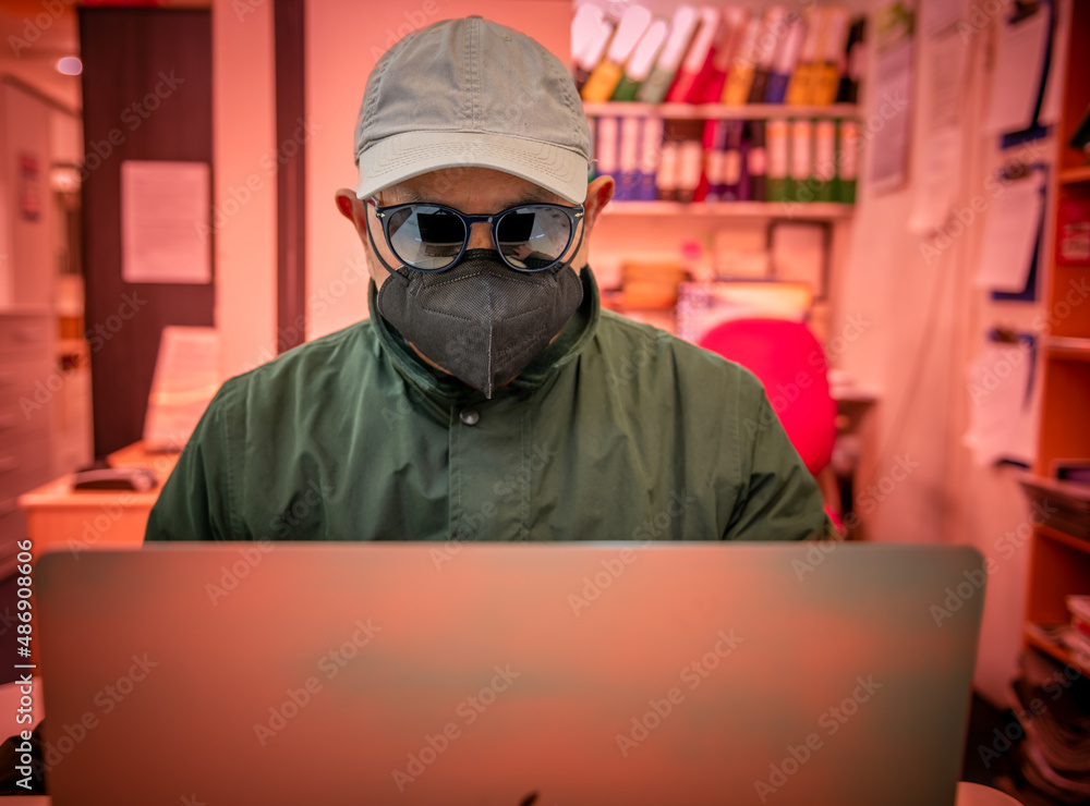 A person wearing sunglasses, a cap and face covering using a laptop. Cyber threat, security and financial scams concept. A person wearing sunglasses, a cap and face covering using a laptop. Cyber threat, security and financial scams concept.