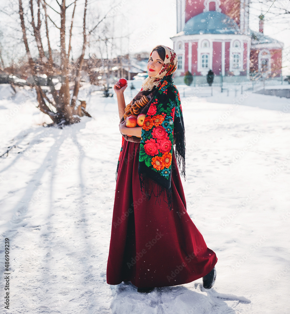 Outdoors lifestyle portrait of pretty young woman in a traditional Russian headscarf holding in hands red apples and bagels on winter background. Maslenitsa festival. Wearing a Russian folk clothes. 