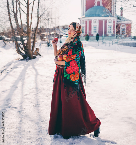 Outdoors lifestyle portrait of pretty young woman in a traditional Russian headscarf holding in hands red apples and bagels on winter background. Maslenitsa festival. Wearing a Russian folk clothes.  © Olesya Kuprina