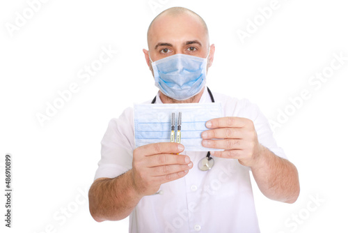 Middle-aged doctor in a white robe with a stethoscope around his neck on a white background holding two covid syringes and a mask