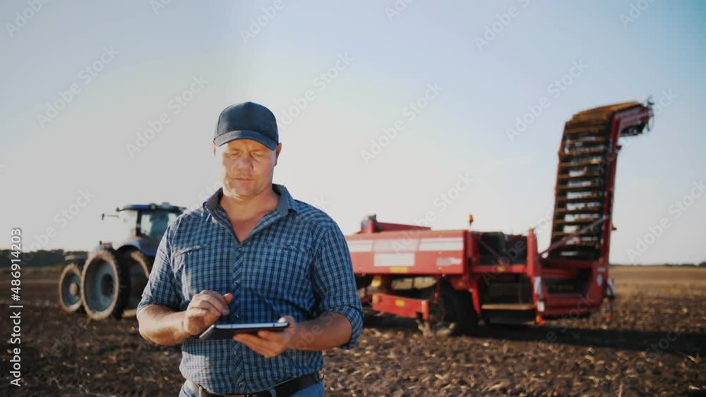 farmer using digital tablet. on farm field, on farm machinery backdrop. tractor with potato harvester. smart farming. farming technologies. potato harvesting. farmer using digital tablet. on farm field, on farm machinery backdrop. tractor with potato harvester. smart farming. farming technologies. potato harvesting.
