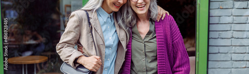 Pretty senior Asian lady hugs grey haired friend in purple jacket standing near cafe entrence on city street. Long-time friendship relationship photo