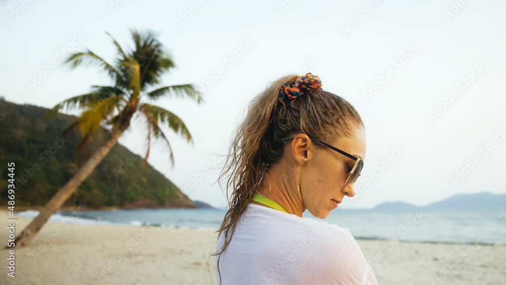 Close up portrait woman in a white tunic shirt on beach, near stormy sea, tilted palm tree. Attractive girl with wet curly hair, in sunglasses. Female tourist walks has fun rest. Blur mist glow view Close up portrait woman in a white tunic shirt on beach, near stormy sea, tilted palm tree. Attractive girl with wet curly hair, in sunglasses. Female tourist walks has fun rest. Blur mist glow view
