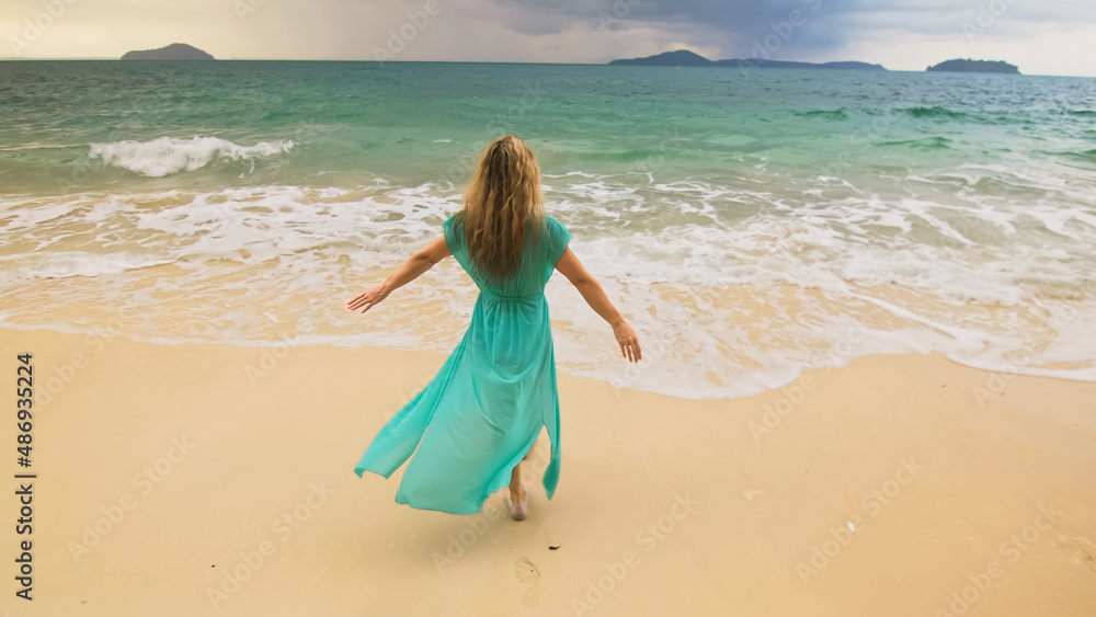 Beautiful long-haired woman walks to the stormy cloudy ocean on sand beach. Girl in blue swimsuit dress tunic. Concept rest in sea, tropical resort coastline traveling tourism summer beach holidays Beautiful long-haired woman walks to the stormy cloudy ocean on sand beach. Girl in blue swimsuit dress tunic. Concept rest in sea, tropical resort coastline traveling tourism summer beach holidays