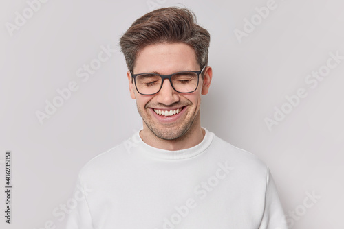Photo of handsome cheerful man with dark hair smiles gladfully with closed eyes expresses positive emotions daydreams about something wears spectacles and casual jumper isolated on whie wall © wayhome.studio 