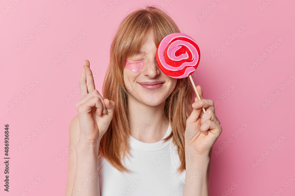 Positive hopeful ginger girl covers eye with sweet lollipop keeps fingers crossed believes in good luck weas casual white t shirt poses over pink background. Lovely redhead female teenager with candy