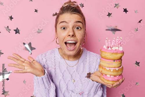 Positive surprised birthday girl feels joyful receives congratuations hlds festive sweet donuts wears casual jumper spends free time on celebration isolated over pink background with confetti around photo