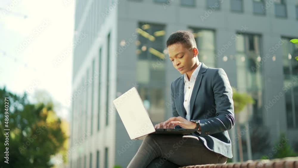 young African American business woman freelancer sitting on bench working with laptop in city park on modern urban street background outdoors downtown Female enjoy outside entrepreneur browsing typing