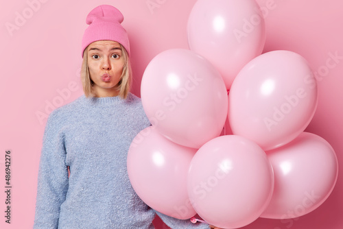 Romantic blonde woman keeps lips rounded wears hat and sweater ready for celebration holds bunch of helium balloons prepares decorations for party isolated over pink background. Holidays concept © wayhome.studio 