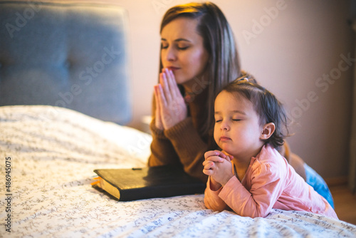 Mother and her daughter reading from bible and praying in their knees near the bed photo