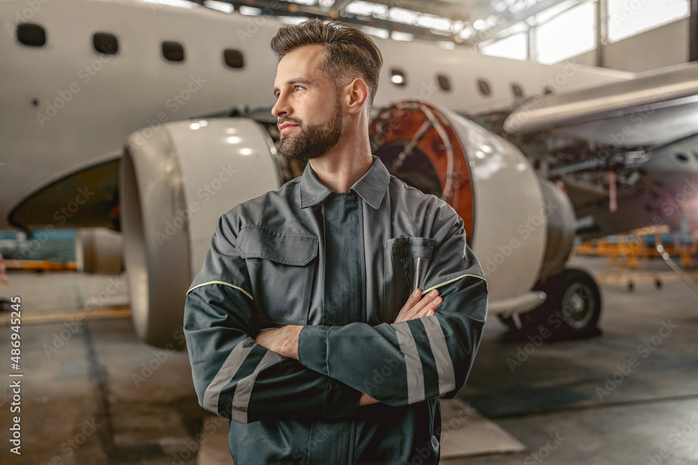 Male aircraft maintenance engineer looking away and keeping arms crossed while standing near passenger plane Male aircraft maintenance engineer looking away and keeping arms crossed while standing near passenger plane