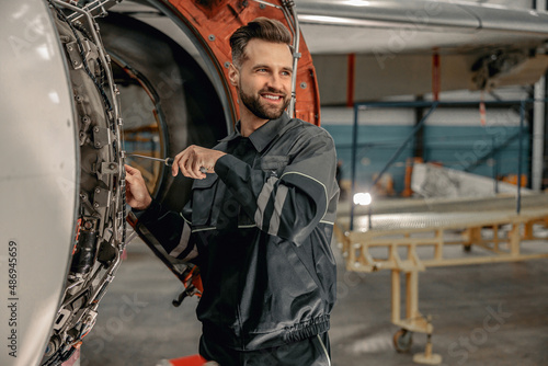 Joyful bearded man maintenance technician using screwdriver and smiling while repairing airplane at repair station