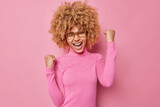 Lucky positive curly haired young woman laughs and winks eye makes triumph gesture wears spectacles and poloneck celebrates triumph isolated over pink background. Yes finally I gained success