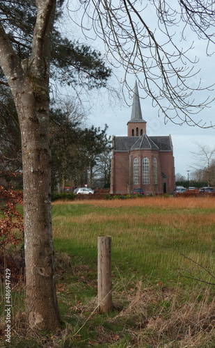 A little village church on a Sunday morning. The mass visitors have parked their car at the church. A bare tree and a pole are in foreground. In between tree and church is a meadow. Laar, Germany photo