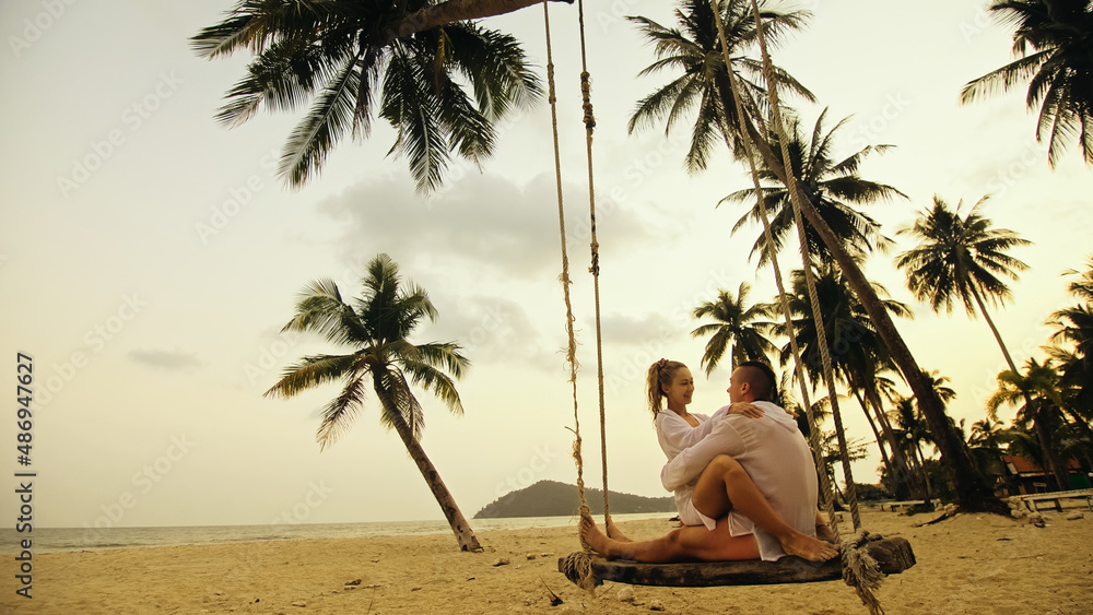 Obraz premium Cute loving couple on the swing on tropical beach. Woman and man in white shirt enjoy gold sunset and life in beaches, palm tree. Evening warm sunset. Summer holiday vacation tropical tourism concept