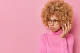 Displeased sullen young woman looks away with upset expression wears transparent glasses casual turtleneck expresses negative emotions poses against pink background copy space for your text.