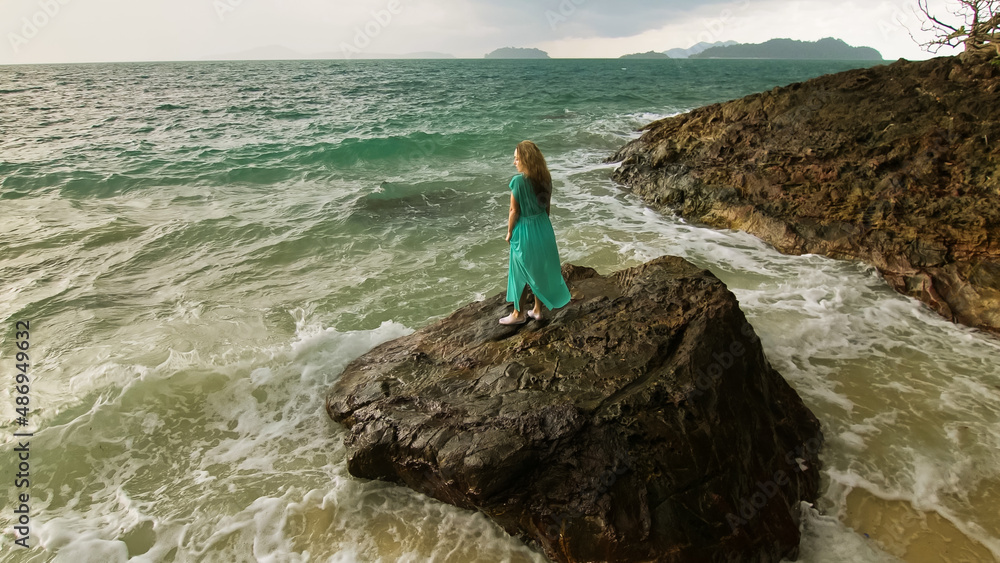 Obraz premium Seductive long-haired woman walks on rock of sea reef stone, stormy cloudy ocean. Woman in blue swimsuit dress tunic. Concept rest in sea, tropical resort coastline traveling tourism summer holidays