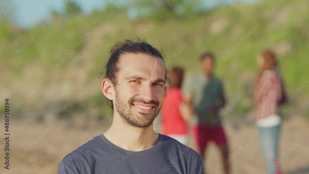 Handsome Hispanic man with curly-haired ponytail and stubble looking and smiling at camera standing on beach, head and shoulders shot. Defocused people talking in background Handsome Hispanic man with curly-haired ponytail and stubble looking and smiling at camera standing on beach, head and shoulders shot. Defocused people talking in background