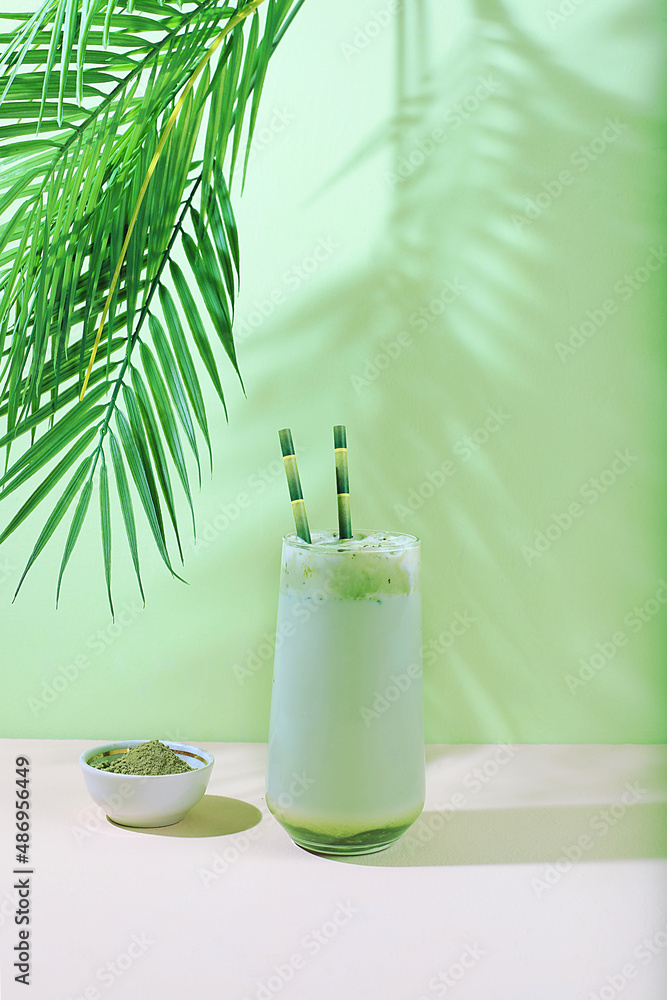 Green tea matcha latte in a glass with bamboo tubes on a modern light background with palm leaves.Traditional Indian drink rich in antioxidants,used for weight loss and treatment,