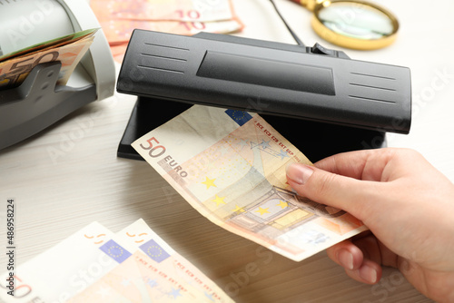 Billede på lærred Woman checking Euro banknote with currency detector at white wooden table, closeup