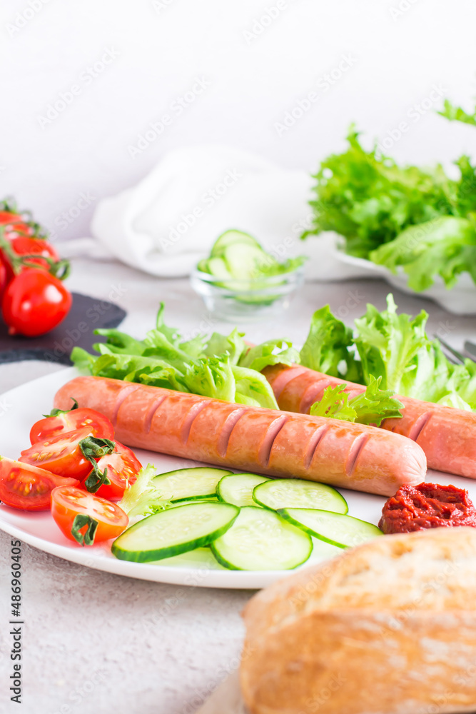 Ready to eat fried sausages, chopped tomatoes and cucumber vegetables on a plate and a bun on the table. Hearty homemade snack. Vertical view Ready to eat fried sausages, chopped tomatoes and cucumber vegetables on a plate and a bun on the table. Hearty homemade snack. Vertical view