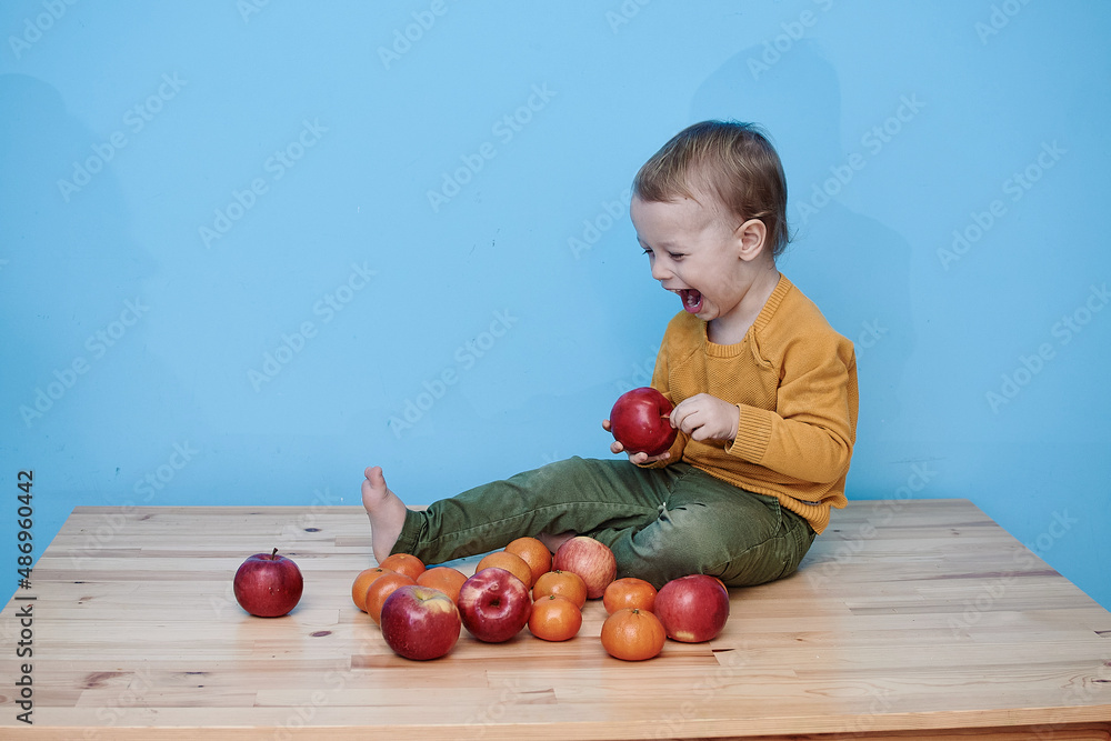 Fototapeta premium 2 year old boy with red apples and tangerines. boy dressed green pants yellow jacket sits advertising vegetables fruits