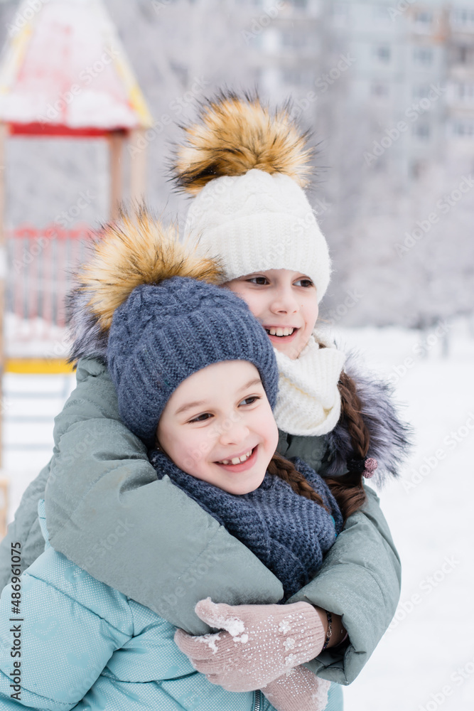 Two smiling girls in warm clothes are hugging in a snowy city. Winter walks, lifestyle. Vertical view