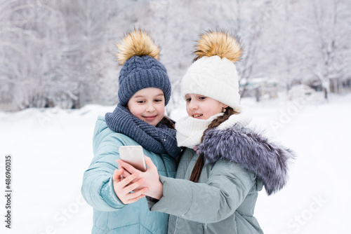 Two smiling girls in warm clothes take a selfie on a smartphone in a snowy winter park. Lifestyle use of technology © elenarui