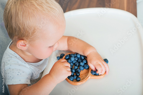 Blonde toddler boy eating Yummy blueberries wooden spoon on highchair close-up and copy space...